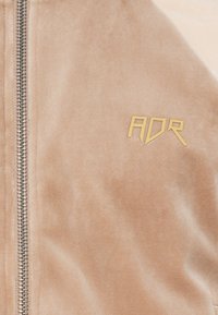 Vaļēta jaka gaiši pelēkā samta ar mīkstu tekstūru. Izkārtota ar sudraba rāvējslēdzēju un zelta "ADR" logotipu priekšā.