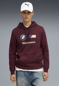 Felpa con cappuccio color bordeaux e tasca frontale, con i loghi BMW e Motorsport. Una maglietta bianca e jeans azzurri chiari completano l'outfit.