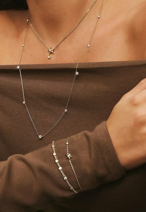 Guldigade lagerhalsband med stjärna och hängande berlock; delikata kedjearmband med cirkulära accenter; uppställda mot en brun tygbakgrund.