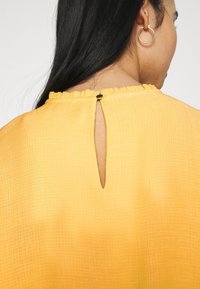 Blusa amarilla con un acabado texturizado, cuello con volantes y abertura en la parte posterior, asegurada con un pequeño botón negro.