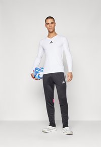 Balta ilga rankovė marškiniai su juodu Adidas logotipu, pilki treningai su rožinėmis detalėmis ir trimis baltais dryžiais, laikantis futbolo kamuolį.