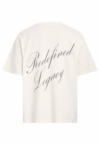 Cremfarbener Baumwoll-T-Shirt mit kurzen Ärmeln. Auf der Rückseite befindet sich der Text "Redefined Legacy" in einer eleganten schwarzen Schrift.