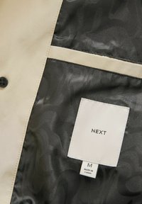 Tissu beige avec doublure à motifs noirs, comportant une étiquette indiquant "NEXT" et "FABRIQUÉ EN CHINE," taille moyenne, détail de bouton visible.
