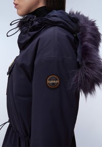Marinblå pufferjacka med pälsfodrad huva, snörning och dragkedjedetaljer, med en cirkulär logotyppatch på vänster ärm.