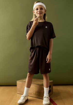 Jonge atleet in donkere sportkleding en een witte hoofdband staat op een houten doos, met een fluitje in de hand, tegen een groene achtergrond.