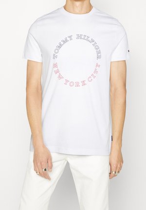 Osoba ubrana w biały T-shirt z krótkim rękawem, z napisem "Tommy Hilfiger" i "New York City" ułożonym w okrągły wzór na piersi.