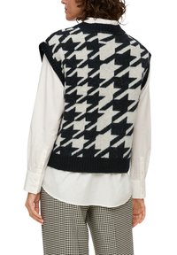 Gilet en motif pied-de-coq noir et blanc, en laine tricotée, avec un col et un ourlet côtelés, porté par-dessus une chemise blanche à col boutonné.