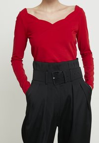 Rotes Langarmoberteil mit weitem V-Ausschnitt, kombiniert mit hochtaillierten schwarzen Hosen, die einen breiten Gürtel und ausgeprägte Falten aufweisen.
