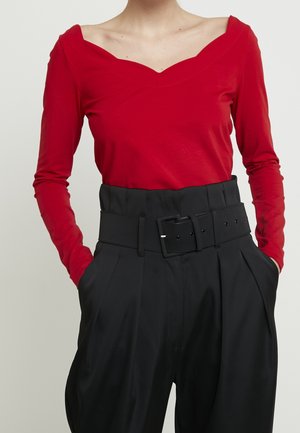 Maglietta rossa a maniche lunghe con scollo a V ampio, abbinata a pantaloni neri a vita alta con una cintura spessa e pieghe evidenti.