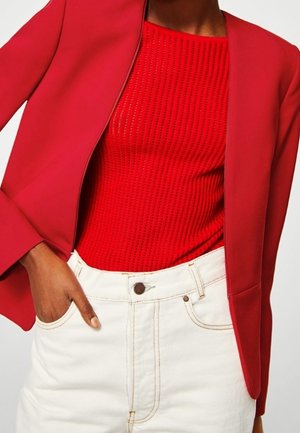 Person trägt ein rotes, strukturiertes Strickoberteil unter einem roten Blazer, kombiniert mit hoch taillierten weißen Denim-Hosen, eine Hand in der Tasche.