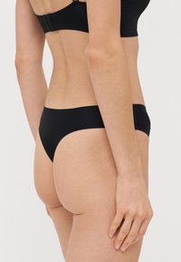 Μαύρο κάτω μέρος μπικίνι με λεία υφή, χαμηλή κοπή και μινιμαλιστικό σχέδιο, απεικονιζόμενο από το πίσω μέρος σε φωτεινό φόντο.