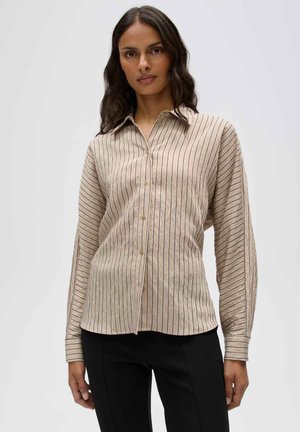 Femme portant une chemise beige à manches longues boutonnée avec de fines rayures verticales noires et un pantalon noir sur un fond uni.