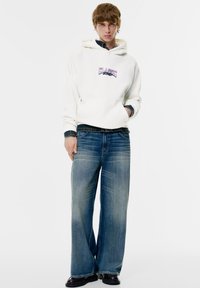 Witte hoodie met "CLASSIC" opdruk; donkerdenim overhemd eronder, wijde blauwe jeans met vervaging, zwarte laarzen. Eenvoudige, casual look.