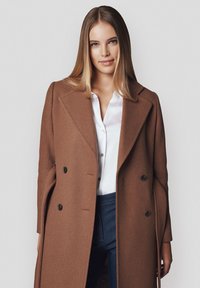 Brązowy płaszcz z mieszanki wełny z podwójnym zapięciem, czarnymi guzikami, klapami z wywinięciem i eleganckim krojem, noszony na białej koszuli.