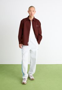 Maroon button-up shirt, white tank top, white pants with blue side stripes, and beige sneakers. Model stands against a light background.