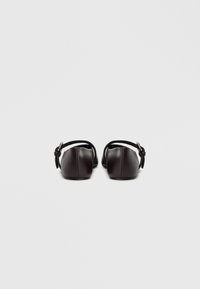 Zapatos planos de cuero marrón con punta redondeada y correas ajustables en el tobillo, que presentan una textura lisa y un diseño minimalista.