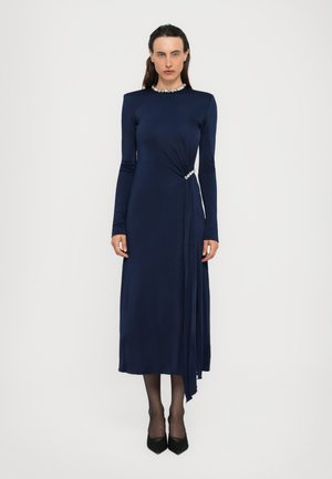 Una mujer se encuentra de pie vistiendo un largo vestido azul marino con mangas largas y detalles de strass en el escote y la cintura, combinado con tacones negros y medias.