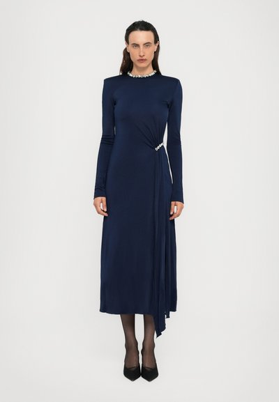 Eine Frau steht in einem langen navyblauen Kleid mit langen Ärmeln und Strassdetails am Ausschnitt und der Taille, kombiniert mit schwarzen High Heels und Strumpfhosen.