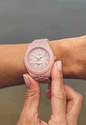 Hellrosa Kunststoffuhr mit strukturiertem Armband und geriffelter Lünette. Roségoldene Stundenmarkierungen und Zeiger, Markenname "Timex" auf dem Zifferblatt angezeigt.