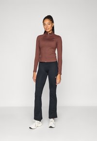 Εφαρμοστή μπλούζα μακρυμάνικη σε σκούρο καφέ ύφασμα με ψηλό γιακά και φερμουάρ μπροστά, συνδυασμένη με μαύρο παντελόνι εφαρμοστό και λευκά αθλητικά παπούτσια.