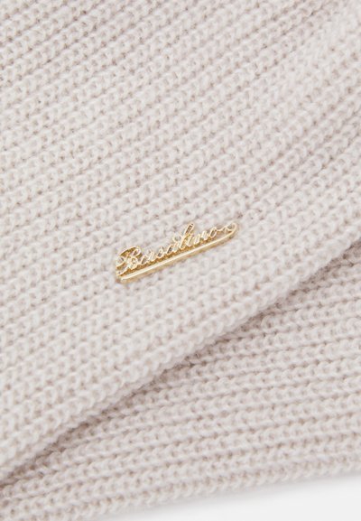 Cream knit fabric with a textured pattern, featuring a gold metal logo plate. The fabric is soft and slightly ribbed.