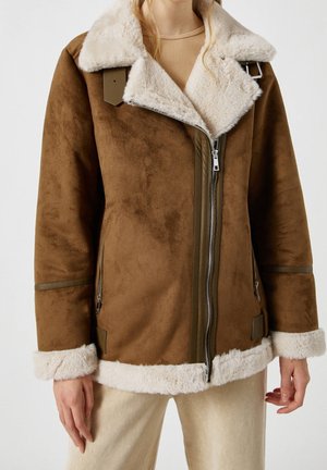 Veste en daim marron avec col, poignets et ourlet en shearling blanc, portée sur un haut côtelé beige et un pantalon clair, fermeture éclair frontale fermée.