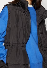 Czarna, pikowana kamizelka z wysokim kołnierzem, z suwakiem i elastycznym paskiem w talii, noszona na niebieskiej bluzie. Gładki, błyszczący materiał.