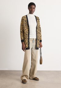 Kardigan z vzorcem leoparda s črnim robom, nošen preko belega zgornjega dela, v kombinaciji s bež širokimi hlačami ter usklajenimi čevlji in torbico z vzorcem leoparda.