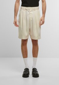 Shorts plissés couleur crème avec une texture lisse, dotés d'une fermeture à bouton, de poches latérales et d'une coupe décontractée, portés avec des chaussures noires et des chaussettes blanches.