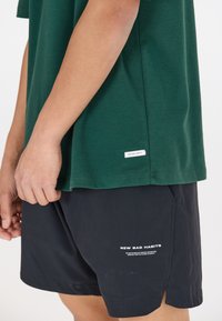 T-paita tummanvihreää puuvillaa, yhdistettynä mustiin shortseihin, joissa on valkoista tekstiä brändäyksenä. T-paidassa on suora helma ja sivumerkintä.