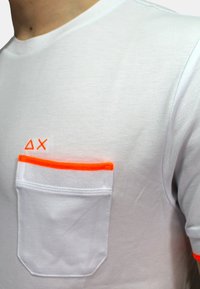 T-shirt bianco in cotone con taschino sul petto rifinito in arancione neon e design geometrico ricamato in arancione sul lato sinistro.