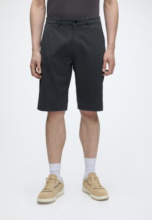 Mann trägt schwarze knielange Shorts, weiße Socken und beige Turnschuhe, steht vor einem schlichten weißen Hintergrund.