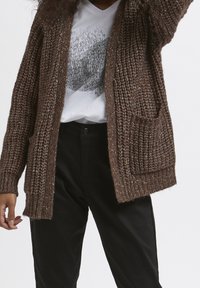 Personne portant un cardigan marron en tricot par-dessus un t-shirt blanc avec un motif graphique et un pantalon noir, debout devant un fond uni.