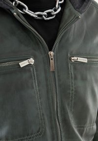 Giacca in pelle verde con due tasche frontali con zip, dettagli in metallo argentato e un cappuccio scuro. Texture liscia con cuciture visibili.