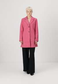 Roze wollen blendjas met een geknipt kraag, twee knopen en een losse pasvorm. Gedragen over zwarte wijde broek, met stijlvolle zwarte schoenen.