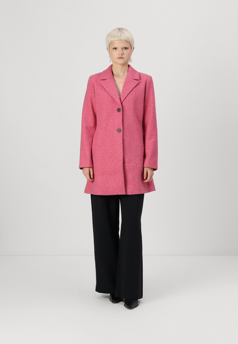 Roze wollen blendjas met een geknipt kraag, twee knopen en een losse pasvorm. Gedragen over zwarte wijde broek, met stijlvolle zwarte schoenen.
