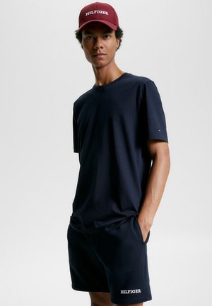 T-shirt in cotone blu navy a maniche corte abbinata a pantaloni corti coordinati. Un cappellino rosso con il logo "HILFIGER" in bianco. Design minimalista.