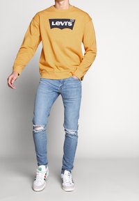 Felpa gialla senape con logo nero Levi's, abbinata a jeans strappati azzurri e sneakers bianche con accenti di colore. Outfit casual.