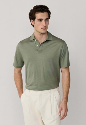 Hombre con polo de manga corta color verde oliva, pantalones blancos y mocasines marrones, de pie con una mano en el bolsillo contra un fondo liso.