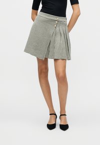 Φούστα με σχέδιο houndstooth και πιέτες, δύο κουμπιά μπροστά και εφαρμοστή μέση σε μαύρο και ελεφαντόδοντο χρώμα, σε συνδυασμό με μαύρες γόβες.