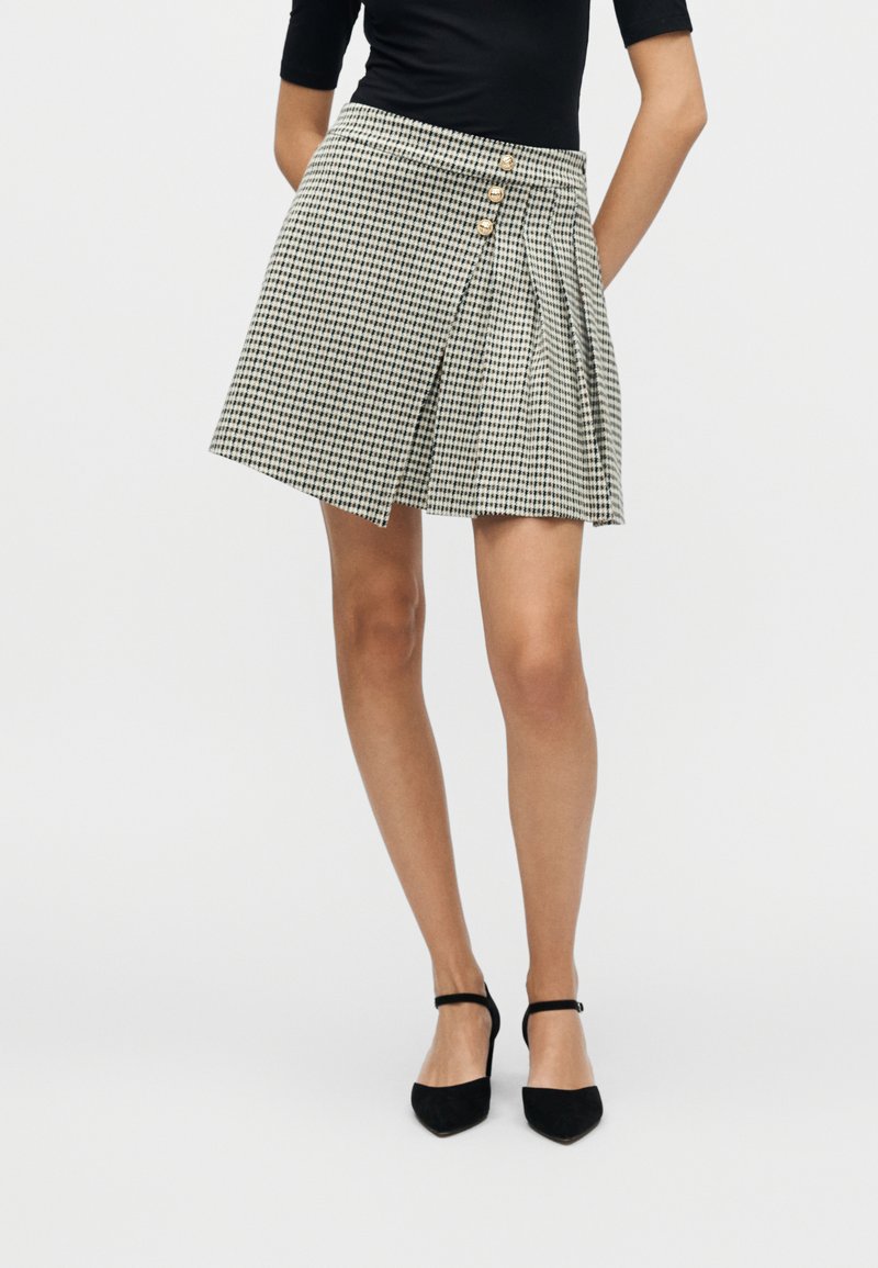 Φούστα με σχέδιο houndstooth και πιέτες, δύο κουμπιά μπροστά και εφαρμοστή μέση σε μαύρο και ελεφαντόδοντο χρώμα, σε συνδυασμό με μαύρες γόβες.