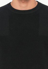 Pulover negru tricotat, cu textură cu nervuri și guler rotund. Materialul este moale, cu un design fără cusături și un model tonal subtil.