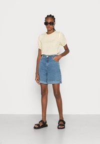 Camisa creme de mangas curtas com padrão texturizado, combinada com calções de ganga azuis de cintura alta e sandálias pretas com tiras grossas.