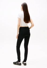 Γυναίκα όρθια με την πλάτη στραμμένη, φοράει λευκό κοντό t-shirt, μαύρο στενό τζιν και μαύρα αθλητικά παπούτσια με λευκές λεπτομέρειες, σε λευκό φόντο.