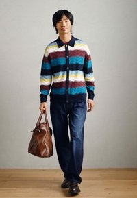 Cardigan tricoté avec un motif zigzag multicolore en bleu, bourgogne, jaune et blanc ; associé à un jean en denim foncé et à un sac en cuir marron.