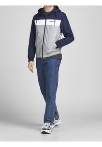 Huvtröja i marinblått, grått och vitt med dragkedja och fickor. Kombinerad med blå jeans och sneakers i flera färger.