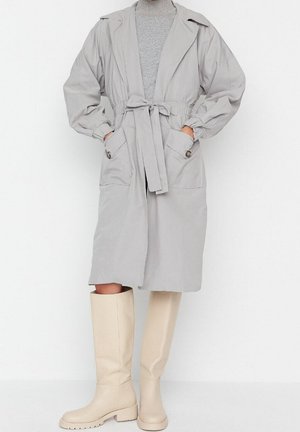 Lichtgrijze trenchcoat met een ceintuur in de taille, elastische manchetten en twee voorzakken. G gecombineerd met kniehoge beige laarzen.