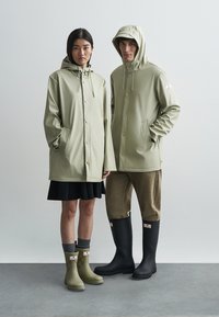 Δύο μοντέλα φοράνε αδιάβροχα μπουφάν ανοιχτού πράσινου χρώματος με κουκούλες, συνδυασμένα με μαύρες και πράσινες λαστιχένιες μπότες, τοποθετημένα μπροστά σε ουδέτερο φόντο.