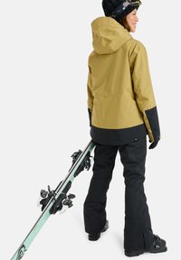 Skijacke in Senfgelb mit schwarzen Akzenten, kombiniert mit schwarzen Hosen. Die Person hält Ski mit weißen Bindungen und trägt Handschuhe sowie eine Mütze.
