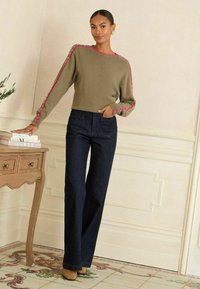 Olívazöld pulóver rózsaszín díszítéssel a ujjakon, sötétkék magas derekú farmernadrággal párosítva. A modell egy faasztal mellett áll.